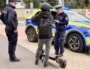Zdjęcie ilustracyjne. małoletni na hulajnodze elektrycznej i policjanci w tle radiowóz.